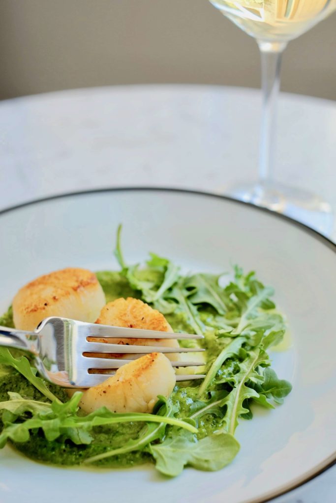 Two pan seared scallops with arugula pesto on a white plate with glass of white wine sitting on a marble table