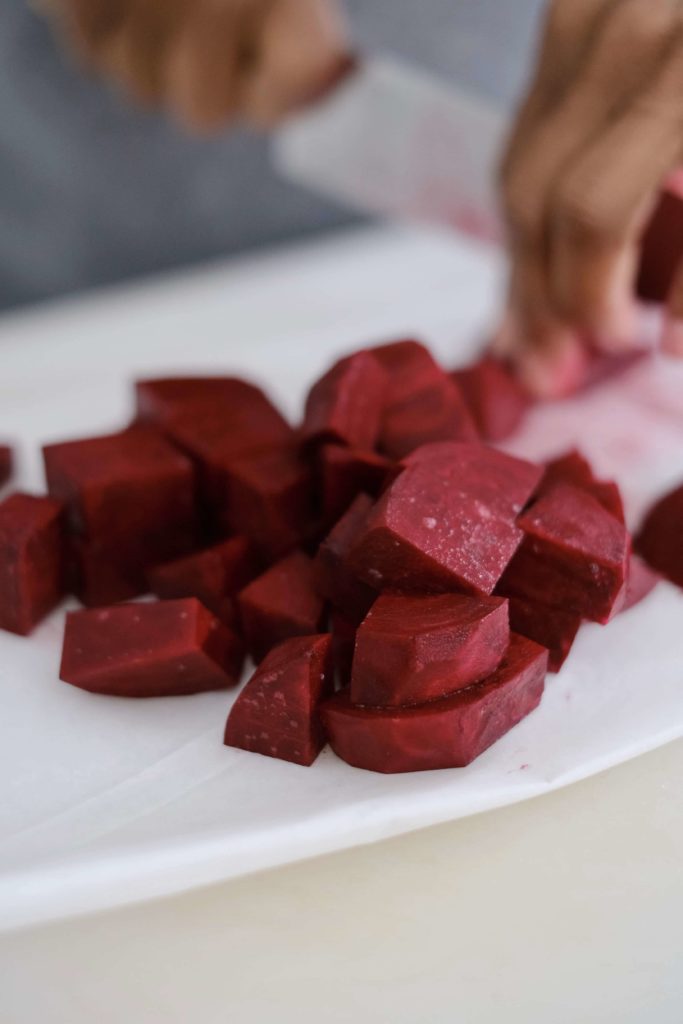 Cubes of raw beets being cut with a knife on a white cutting board