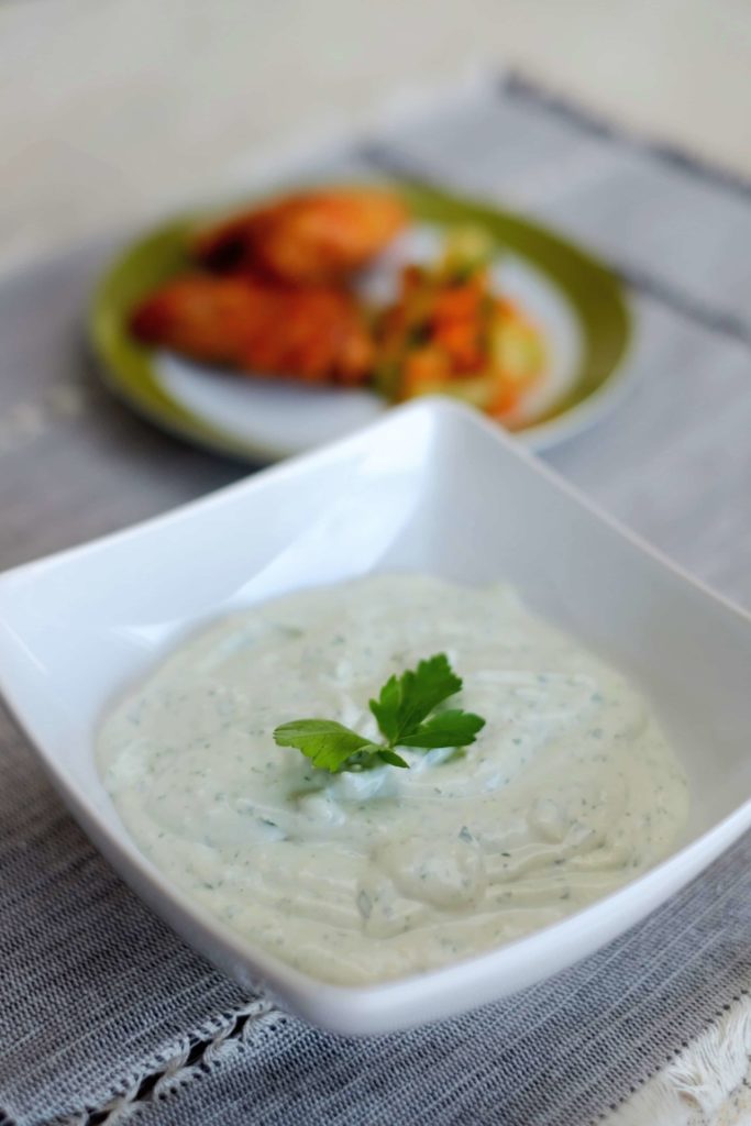 Blue cheese dip in a square white bowl with a parsley sprig on top and chicken wings on a green and white plate in the background,