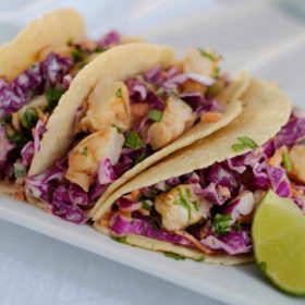 Three grilled shrimp tacos with purple cabbage slaw on a white plate with a lime wedge
