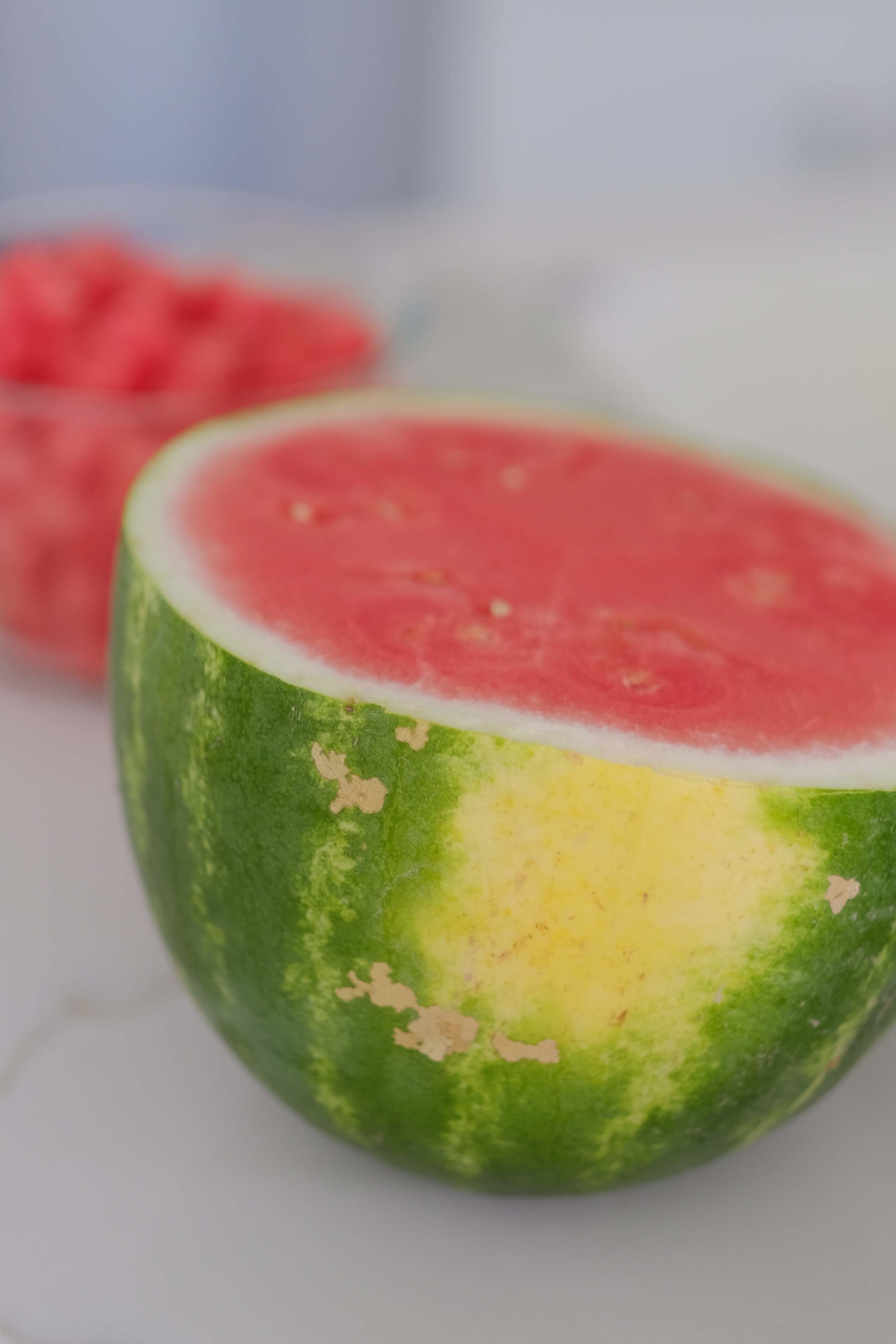 Half a cut seedless watermelon sitting on a white counter with a bowl of cubed watermelon in the background