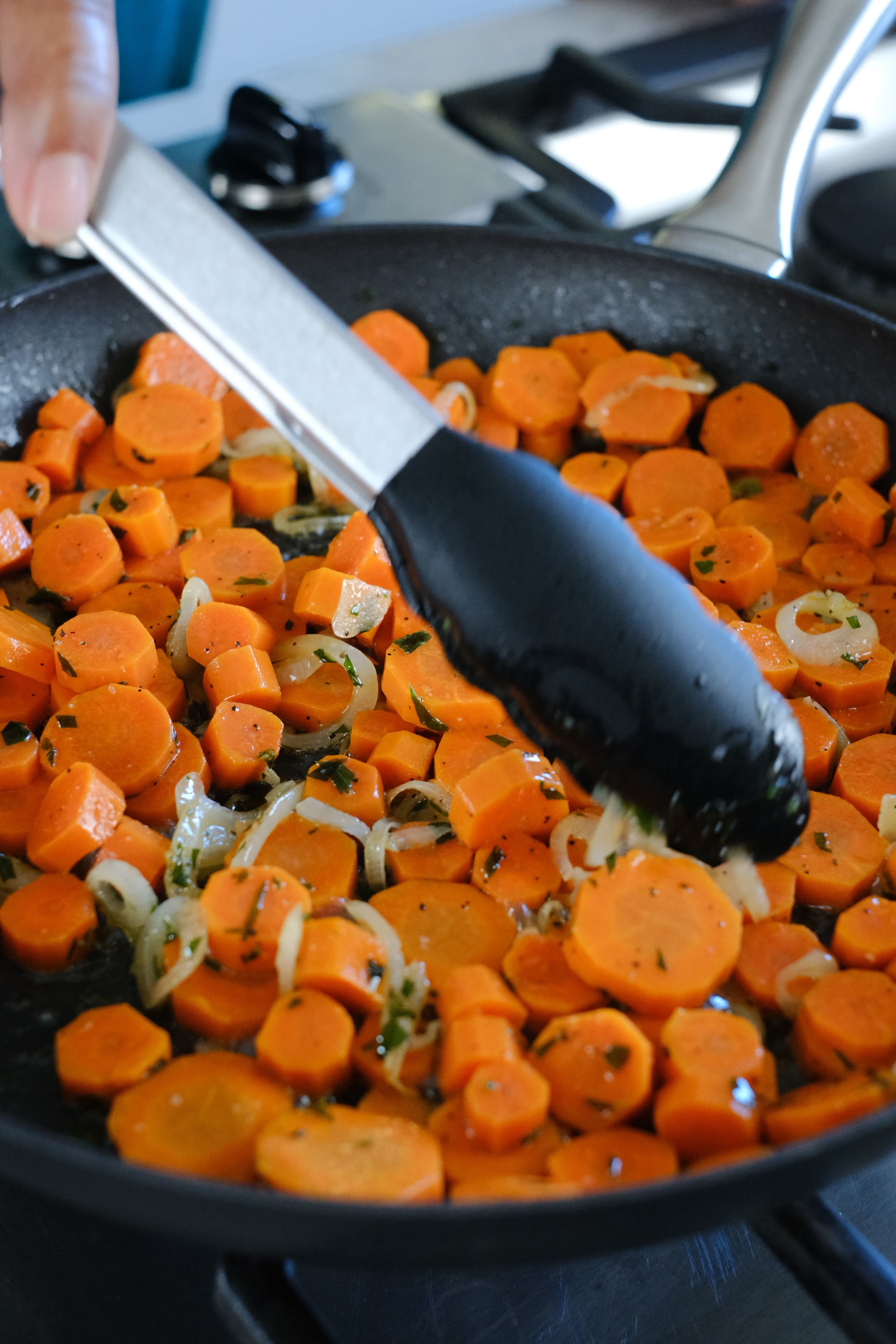 Carrots shallots and tarragon cooking in a black pan with tongs moving the carrots